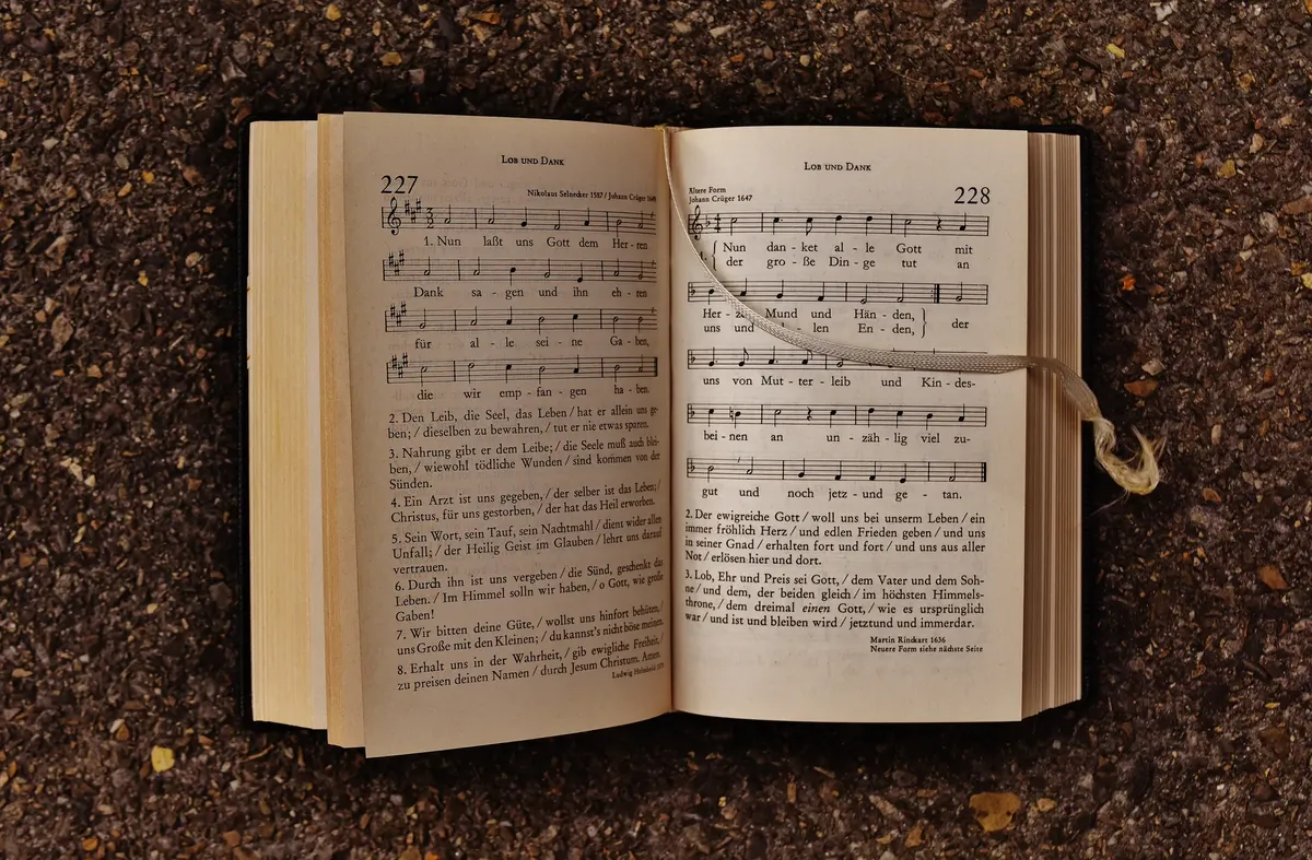 An open hymn book with musical notes laying on earthy ground, featuring sheet music and lyrics.