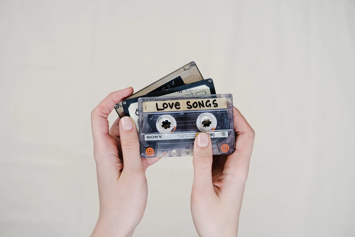 Hands holding cassette tapes labeled 'Love Songs', highlighting 90s nostalgia.