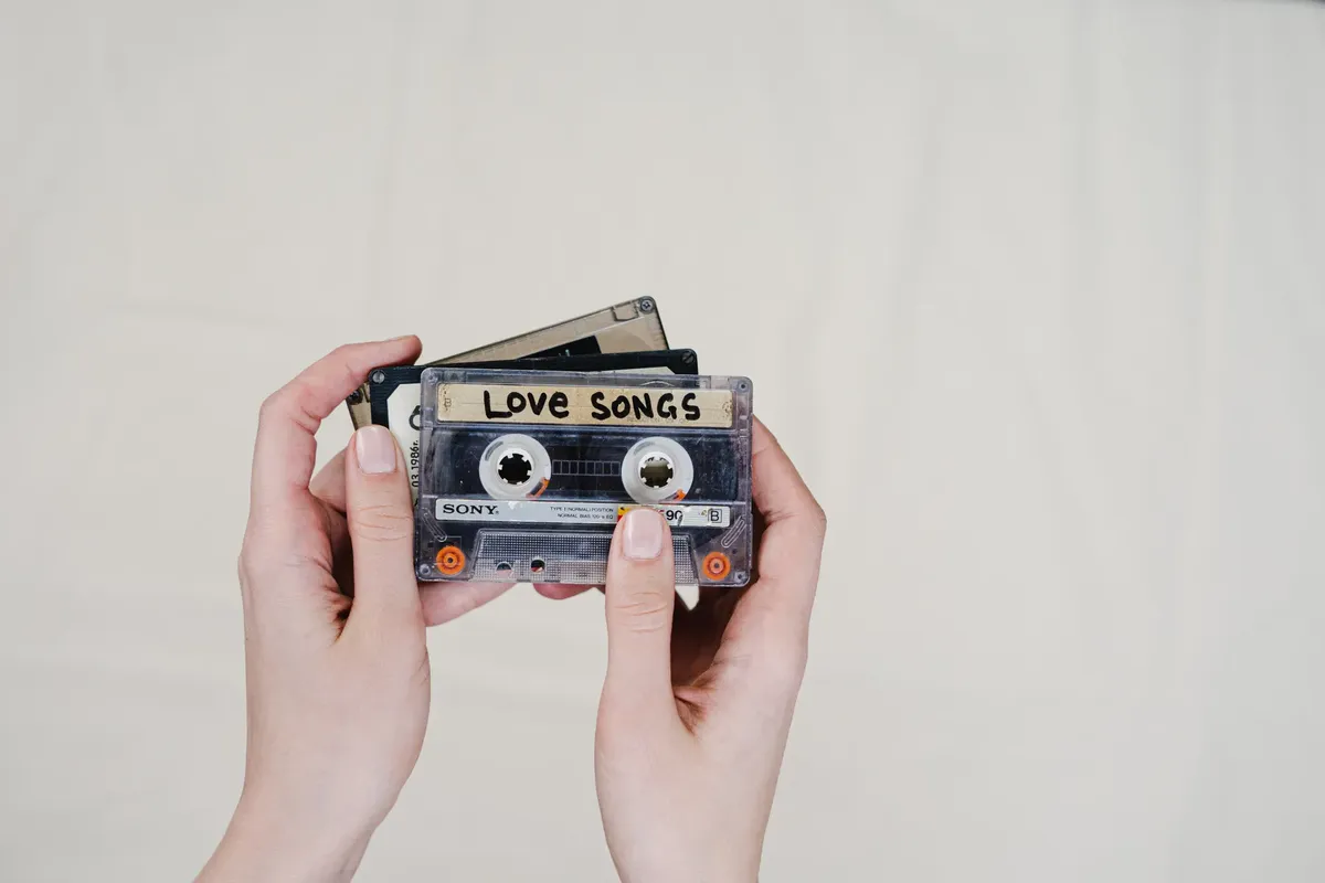 Hands holding vintage cassette tapes, including a 'Love Songs' label, evoking nostalgia.